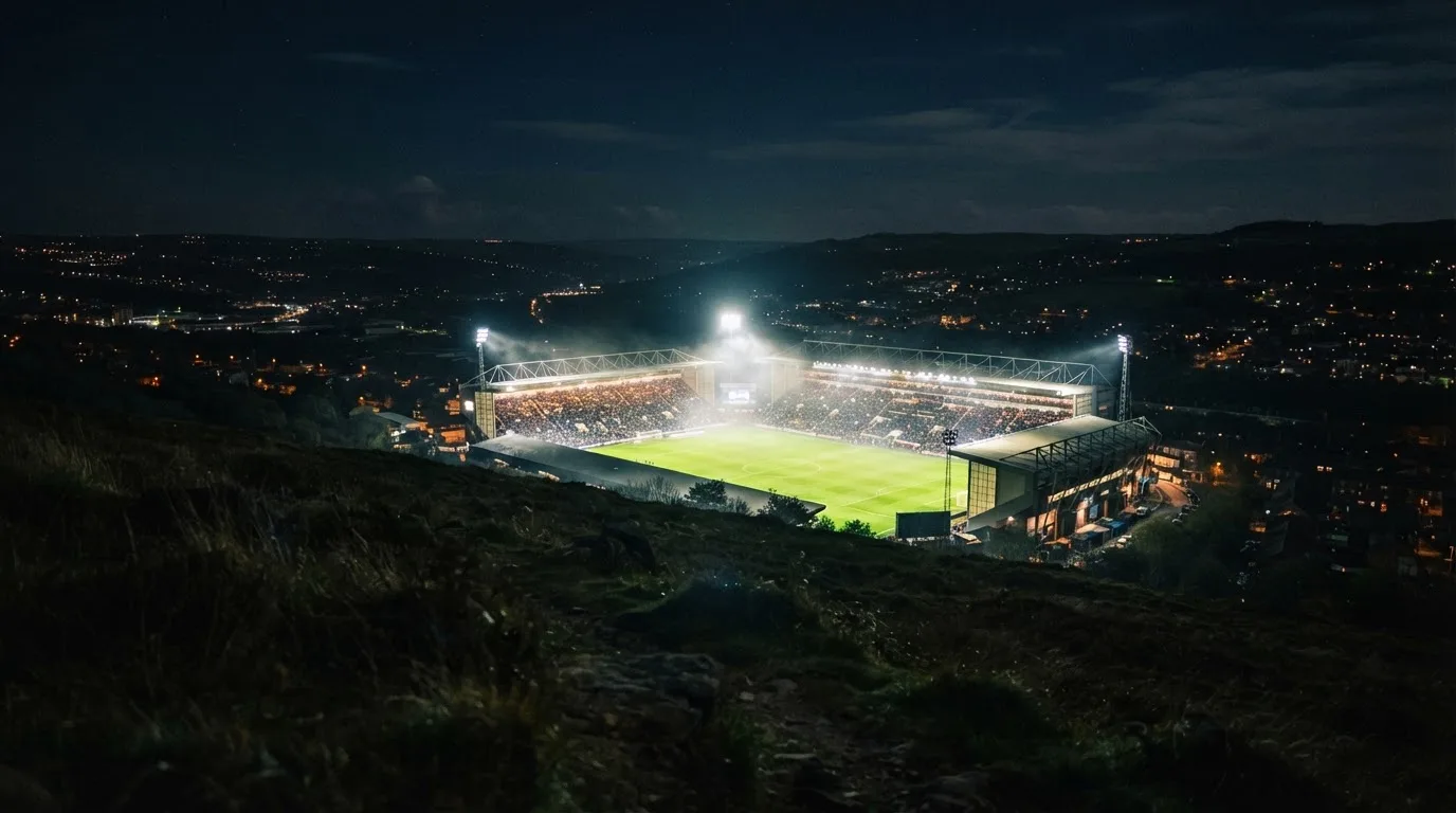Ein Stadion unter Flutlicht bei Nacht, von einem Hügel aus der Ferne betrachtet, mit Dunst über dem Spielfeld.