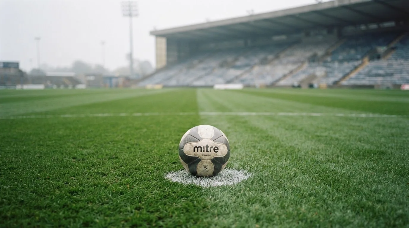 Ein Fußball liegt auf dem Elfmeterpunkt eines leeren Rasenplatzes, im Hintergrund eine Tribüne im Dunst.