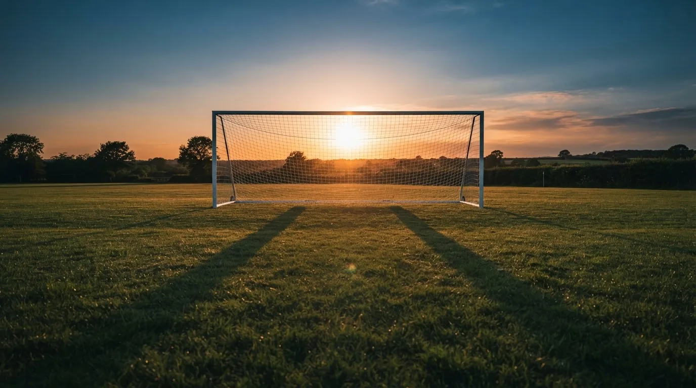 Ein Fußballtor mit Netz bei Sonnenuntergang auf einem leeren Platz, die Schatten der Torpfosten liegen lang auf dem Gras.