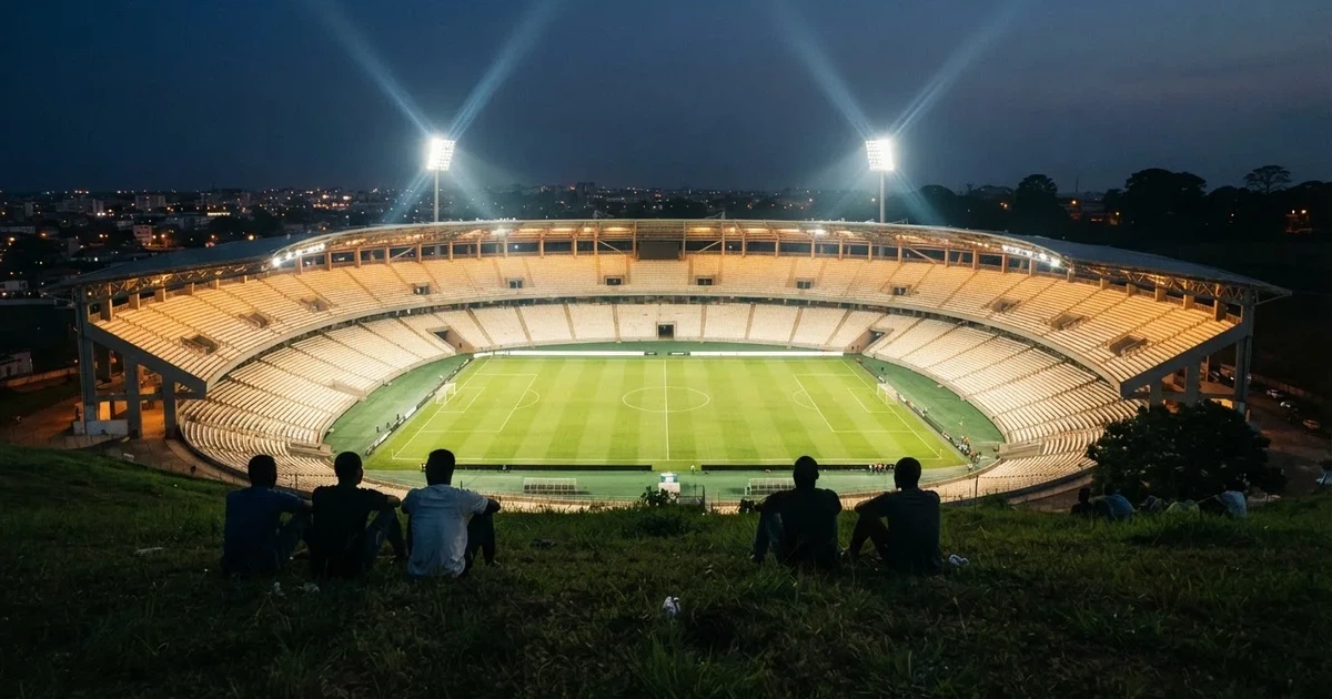 Ein Fußballstadion unter Flutlicht mit hellen Rängen, von einem Hügel aus gesehen, darüber ein dunkelblauer Abendhimmel.