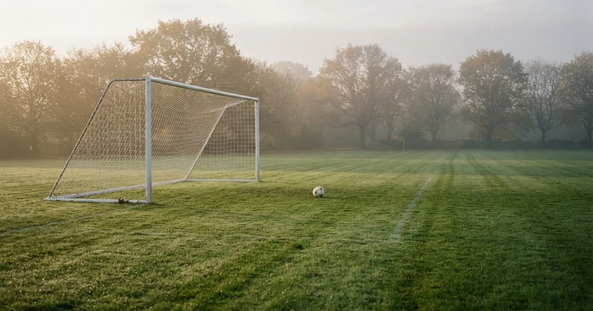 Ein Fußballtor mit gespanntem Netz steht auf einem leeren Rasenplatz, davor ein einzelner Ball, Morgendunst liegt über dem Feld.