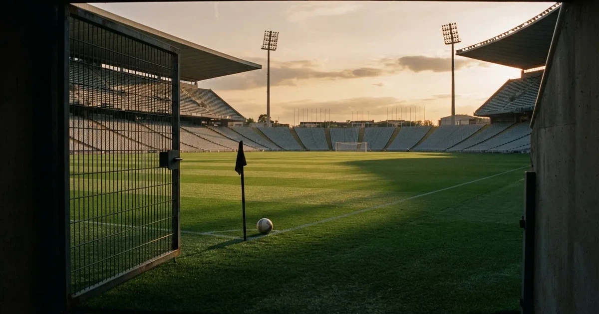 Blick durch ein Stadiontor auf einen grünen Rasen im Abendlicht, ein Ball liegt an der Eckfahne, die Ränge sind leer.