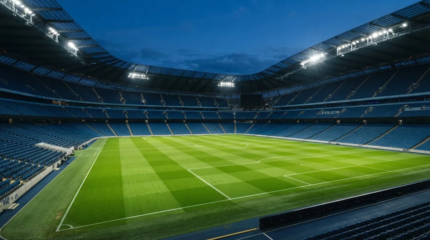 Eine leere Stadiontribüne unter Flutlicht aus der Ferne betrachtet, die Rasenfläche leuchtet grün in der Dämmerung.