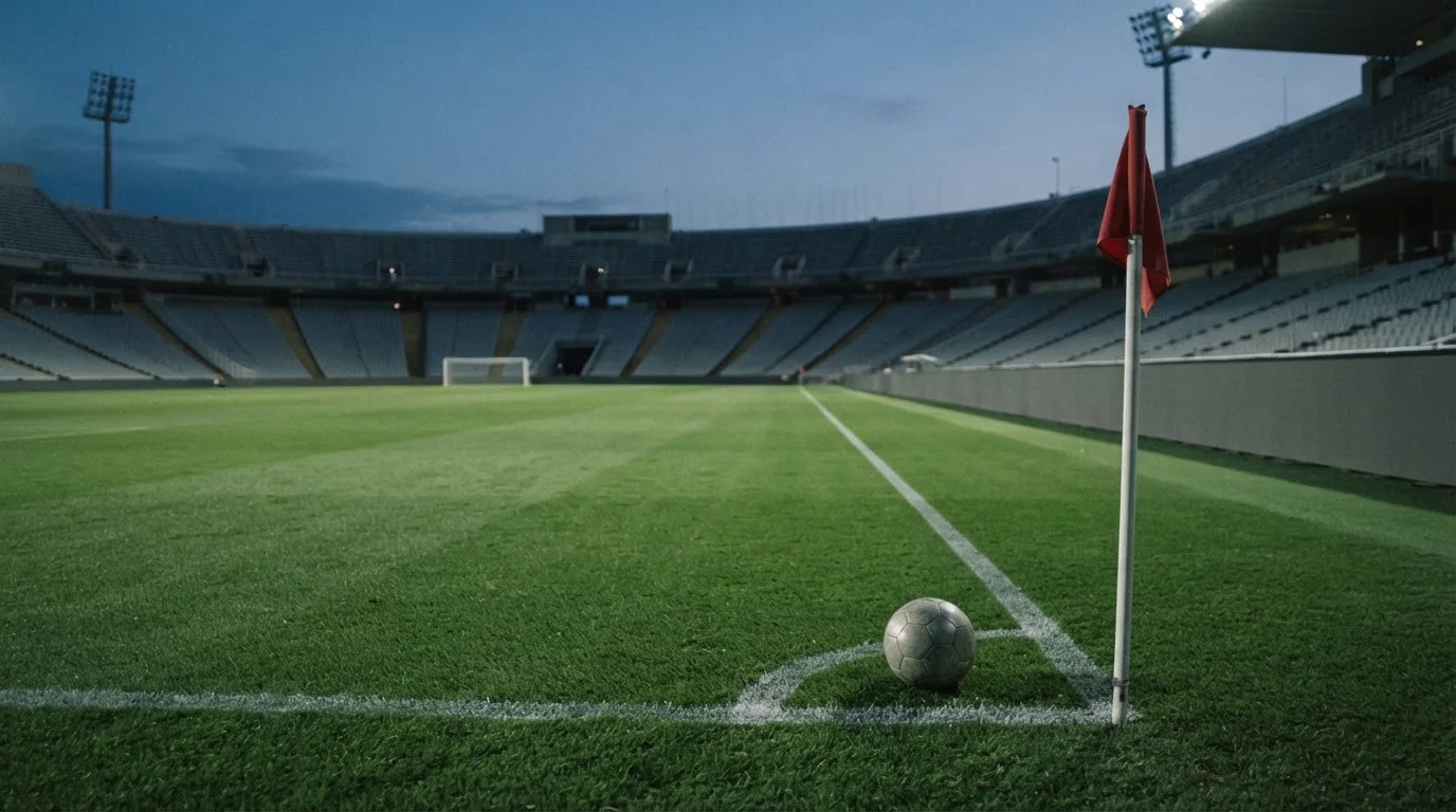 Ein Fußball liegt neben einem Eckfahnen-Pfosten auf einem Rasenplatz, im Hintergrund ein leeres Stadion in der Dämmerung.