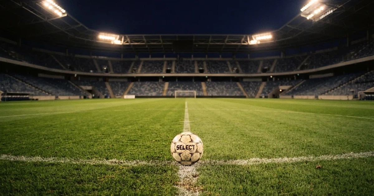 Ein Fußball liegt auf dem Rasen eines leeren Stadions bei Flutlicht, im Hintergrund unscharfe Tribünen ohne erkennbare Beschriftung.