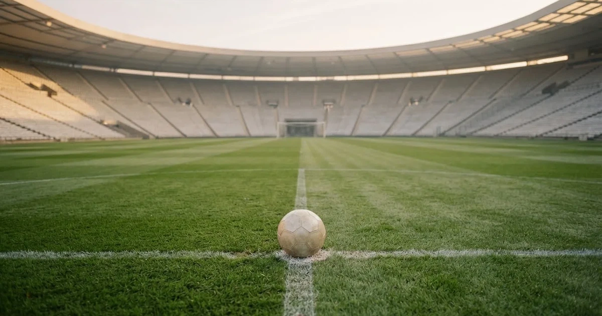 Ein Fußball liegt auf dem Mittelkreis eines Stadionrasens am frühen Morgen, im Hintergrund leere Ränge ohne erkennbare Beschriftung.