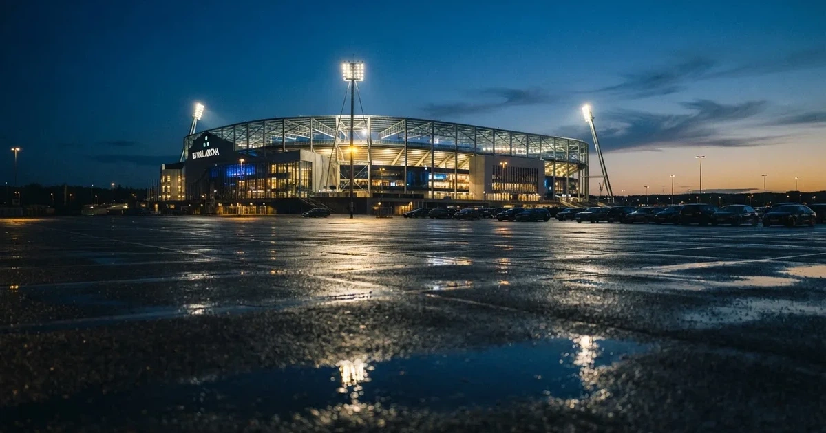 Ein Stadion unter Flutlicht aus großer Entfernung gesehen, davor ein regennasser Parkplatz in der Dämmerung.