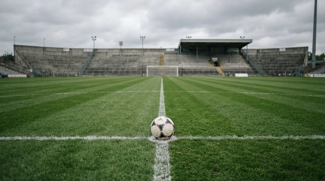 Ein Fußball liegt auf dem Anstoßpunkt eines leeren Rasenplatzes, im Hintergrund eine ruhige Tribüne unter bedecktem Himmel.