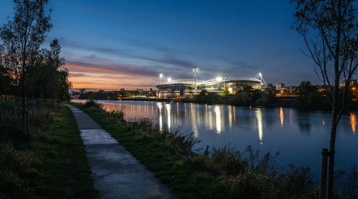 Ein Stadion unter Flutlicht aus der Ferne gesehen, im Vordergrund ein ruhiger Fluss und ein Fußweg.