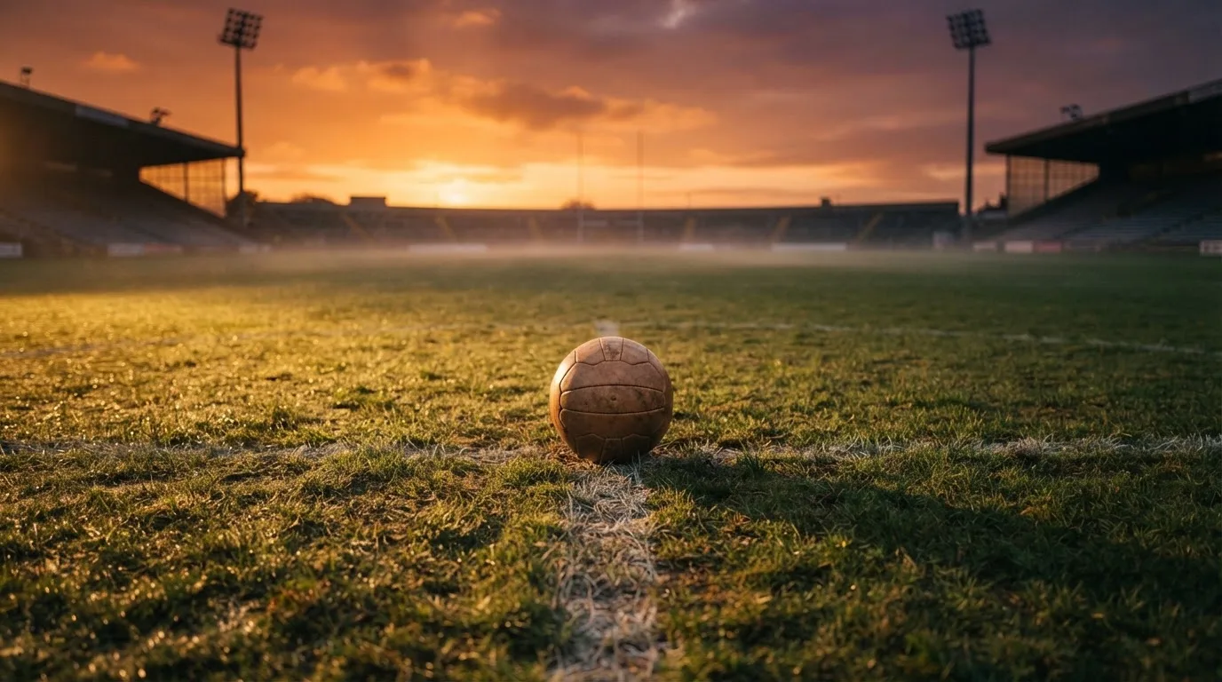 Ein Fußball liegt in der Mitte eines leeren Rasenplatzes, am Horizont färbt sich der Himmel orange.