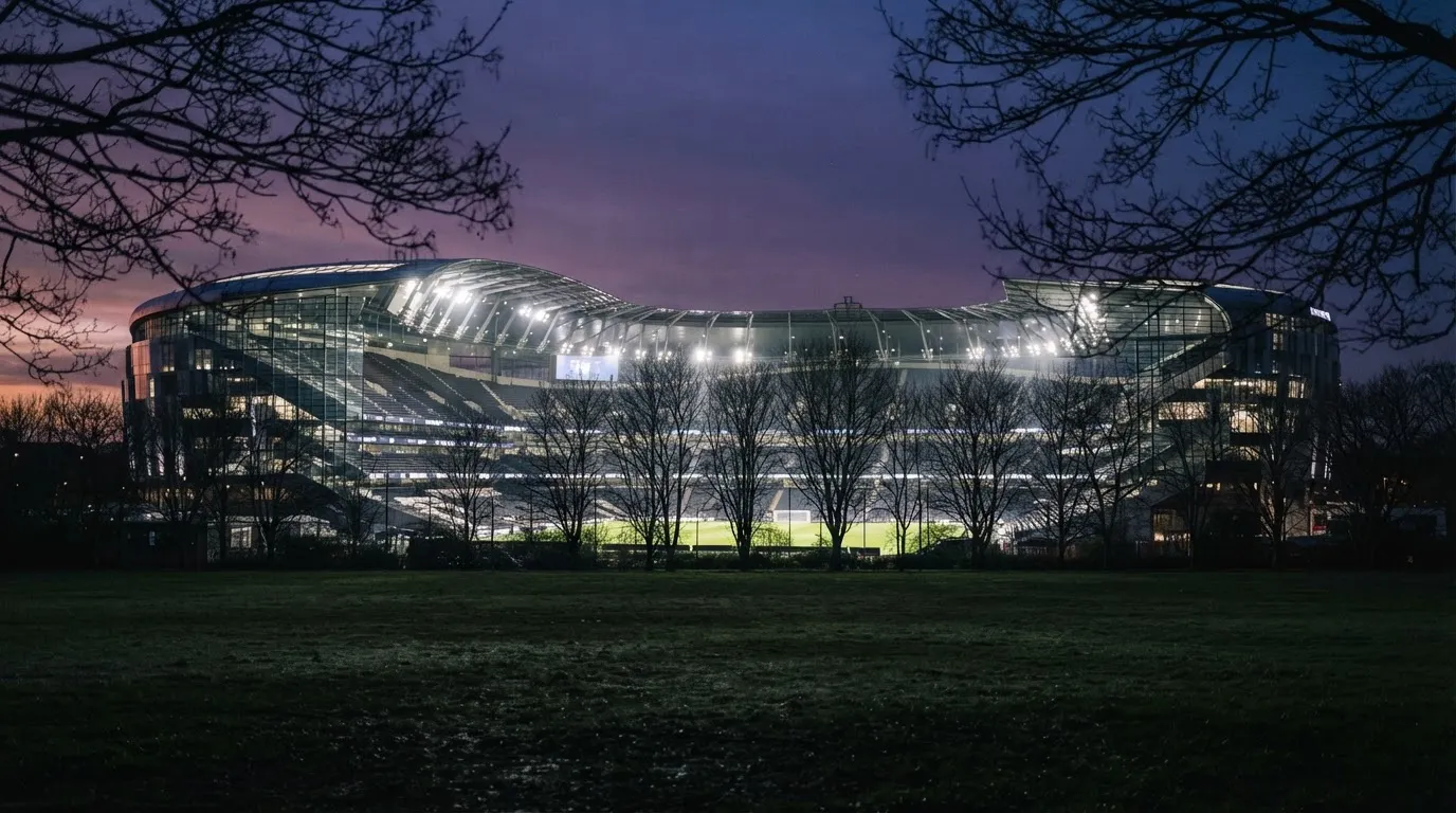 Ein Stadion unter Flutlicht, aus großer Entfernung gesehen, davor eine Reihe kahler Bäume im Dämmerlicht.