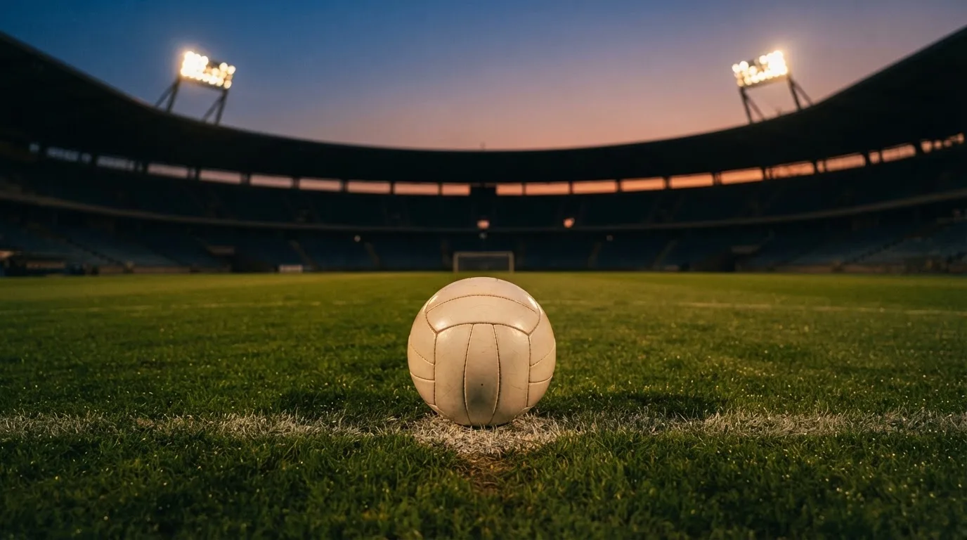 Ein Fußball liegt auf dem Anstoßpunkt eines leeren Rasenplatzes, im Hintergrund steht ein Stadion unter Flutlicht in der Dämmerung.