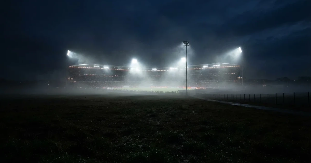 Ein Stadion unter Flutlicht, aus großer Entfernung über eine dunkle Wiese fotografiert, am Himmel zieht Nebel auf.