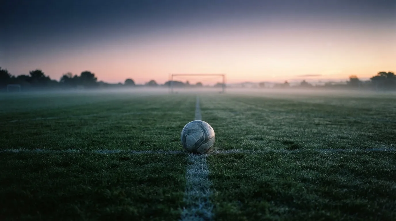 Ein Fußball ruht auf dem Mittelkreis eines leeren Rasenplatzes in der Morgendämmerung, Tau liegt auf dem Gras.