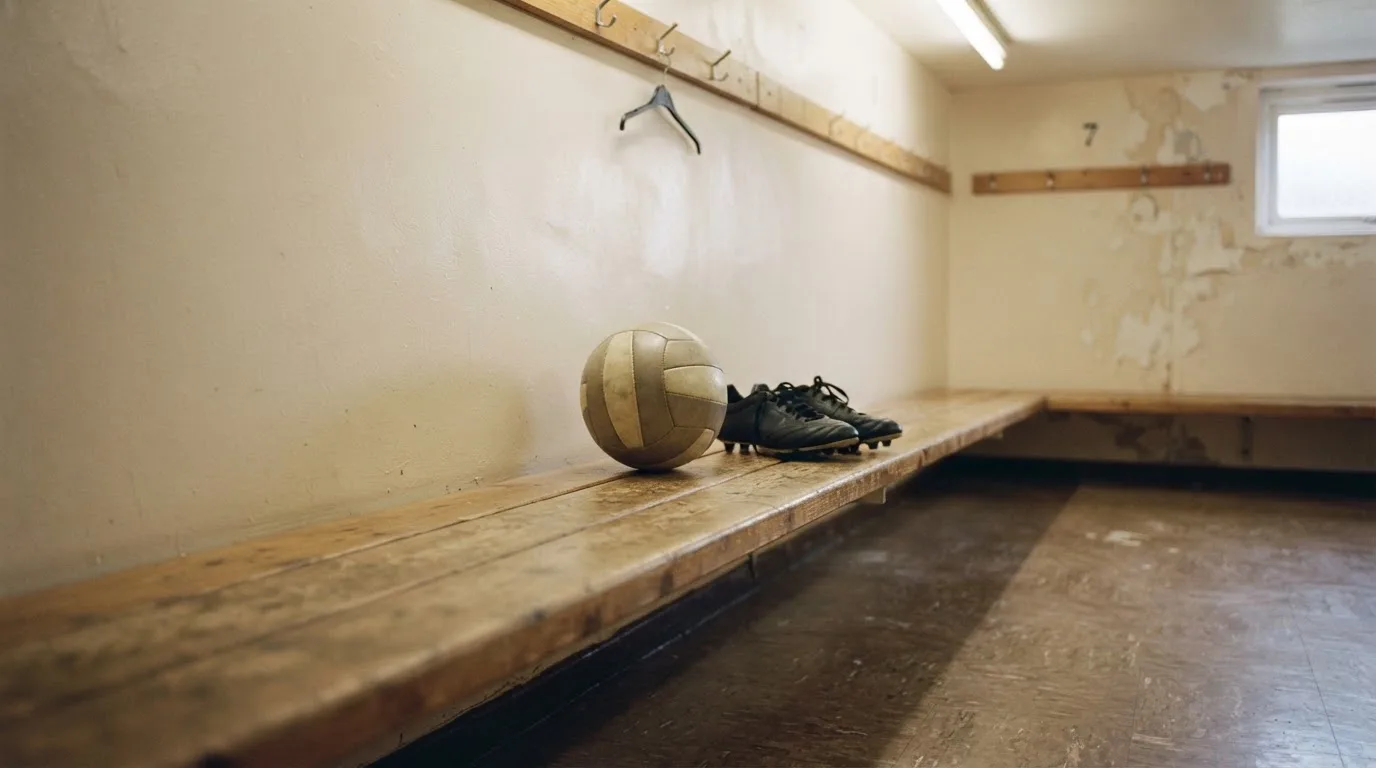 Ein Fußball liegt neben einem Paar Fußballschuhen ohne Markenzeichen auf einer Holzbank in einer leeren Umkleide.