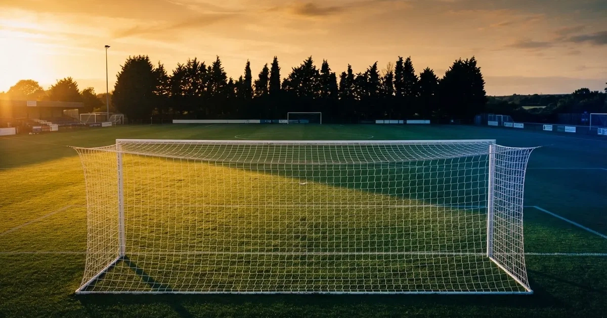 Ein Fußballtor mit Netz auf einem leeren Platz im Abendlicht, der Rasen frisch gemäht, im Hintergrund Zypressen.