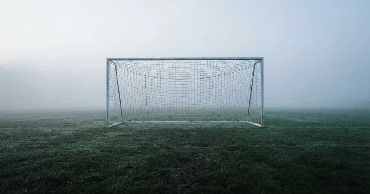 Ein Fußballtor mit Netz steht auf einem leeren Platz im Nebel der Morgendämmerung, der Rasen ist taufeucht.