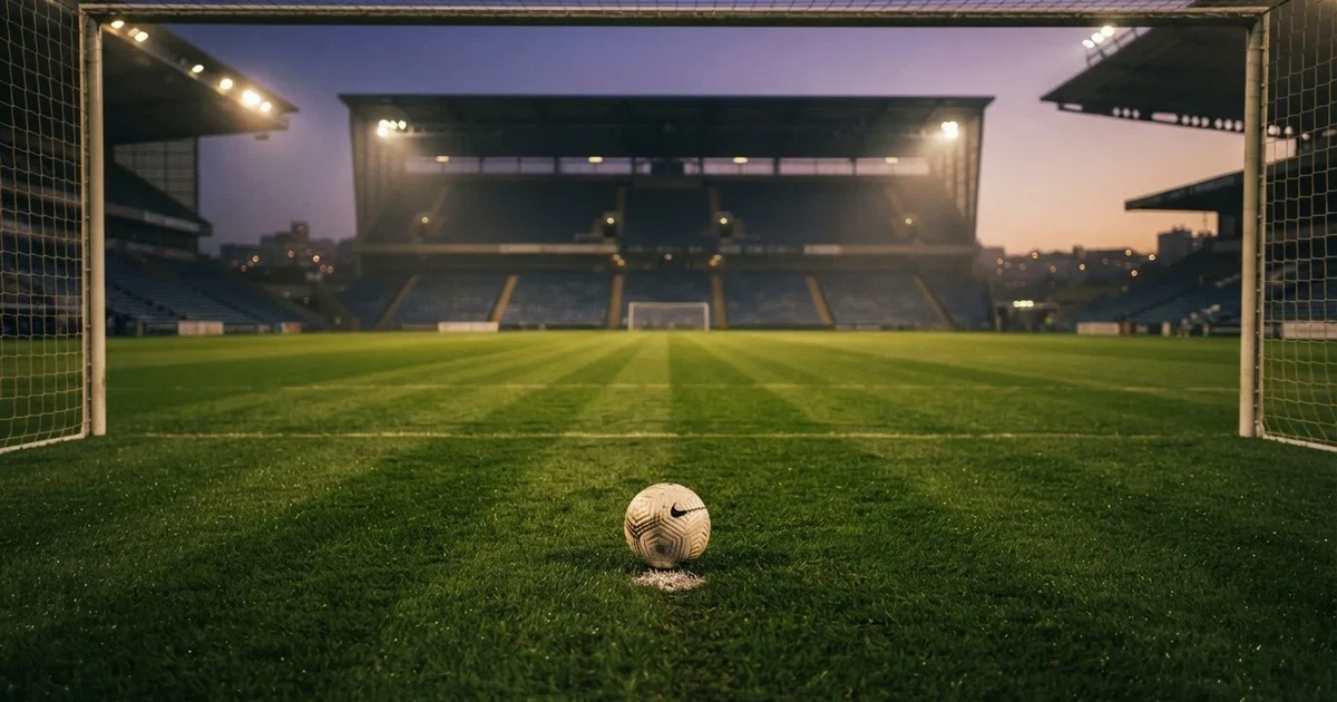 Ein Fußballplatz mit frisch gemähtem Rasen im Abendlicht, ein Ball liegt im Strafraum, im Hintergrund unscharf eine leere Tribüne.