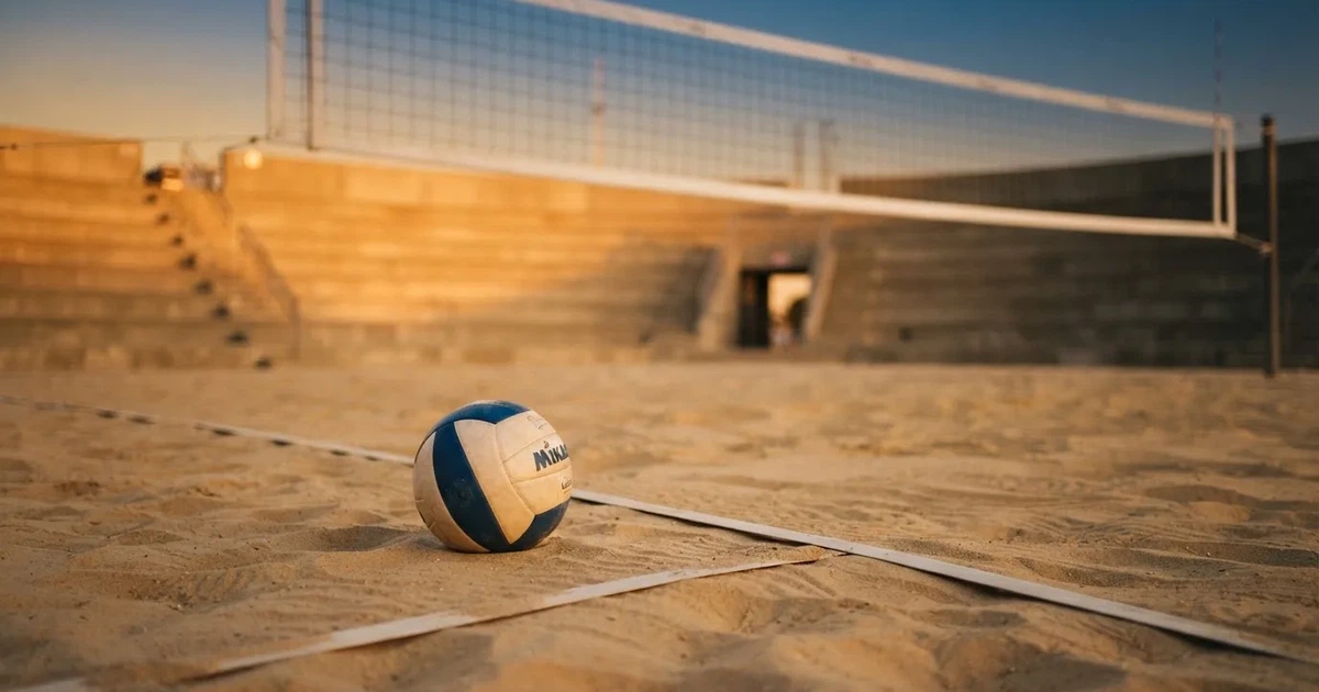 Ein Volleyball liegt auf dem Sand eines Beachvolleyball-Feldes neben dem gespannten Netz, im Hintergrund eine leere Tribüne bei Abendlicht.