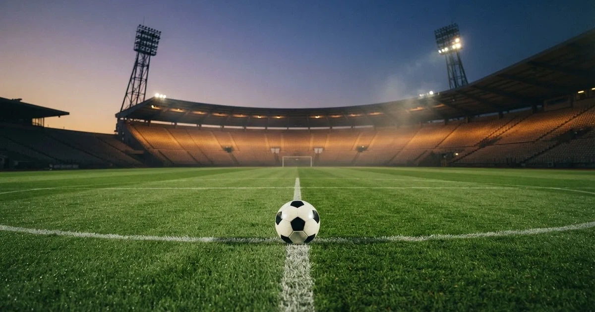 Ein Fußball liegt auf dem Anstoßpunkt eines leeren Rasenplatzes, im Hintergrund eine Tribüne und Flutlichtmasten in der Abenddämmerung.