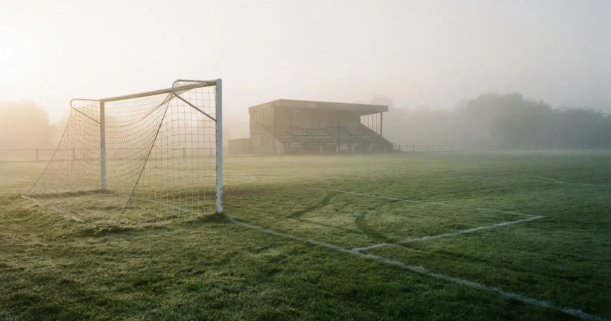 Ein Fußballtor mit Netz steht im Morgennebel auf einem leeren Rasenplatz, im Hintergrund die Silhouette einer kleinen Tribüne.