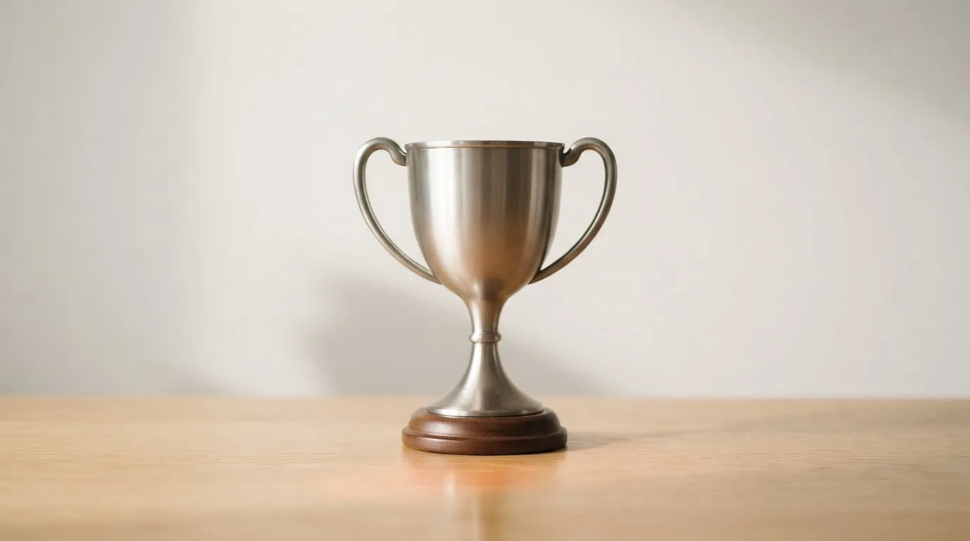 Ein Pokal ohne Gravur steht allein auf einem Holztisch vor einer hellen Wand, davon fällt ein weicher Schatten.