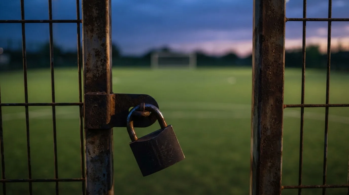 Ein Vorhängeschloss hängt an einem Metalltor in der Dämmerung, im Hintergrund unscharf ein Fußballplatz.
