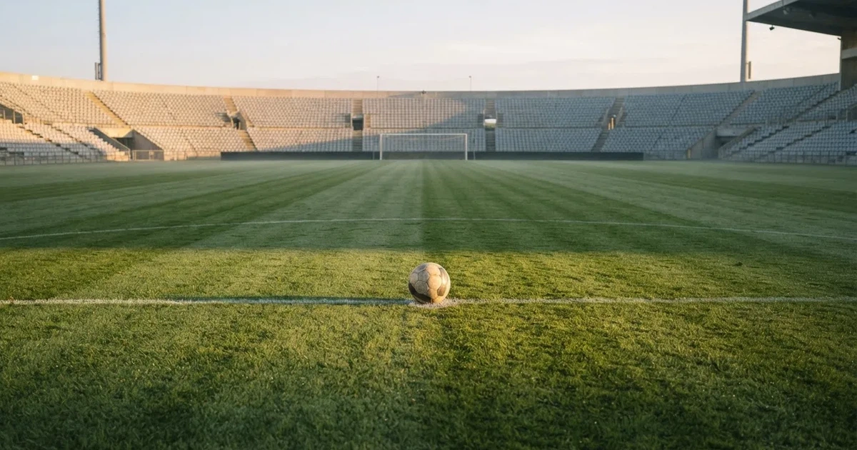 Ein Fußball liegt auf dem Anstoßpunkt eines Rasenplatzes, dahinter leere Tribünen im Licht des frühen Morgens.