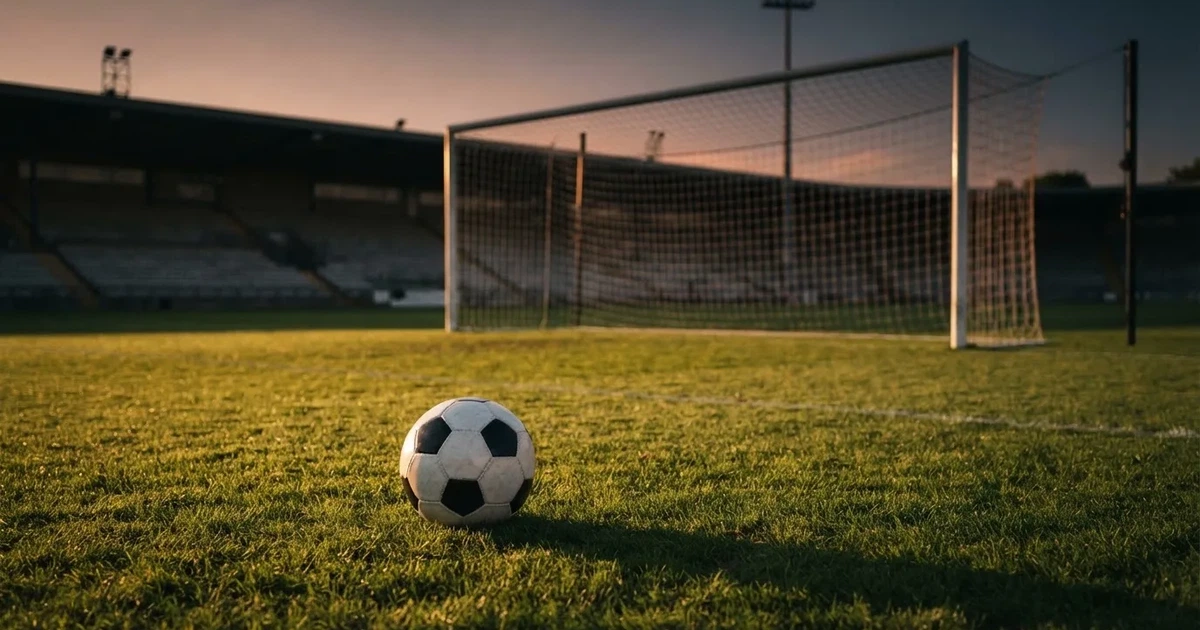 Ein Fußballtor mit Netz im Gegenlicht auf einem leeren Platz am Abend, davor ein Ball auf dem Rasen.