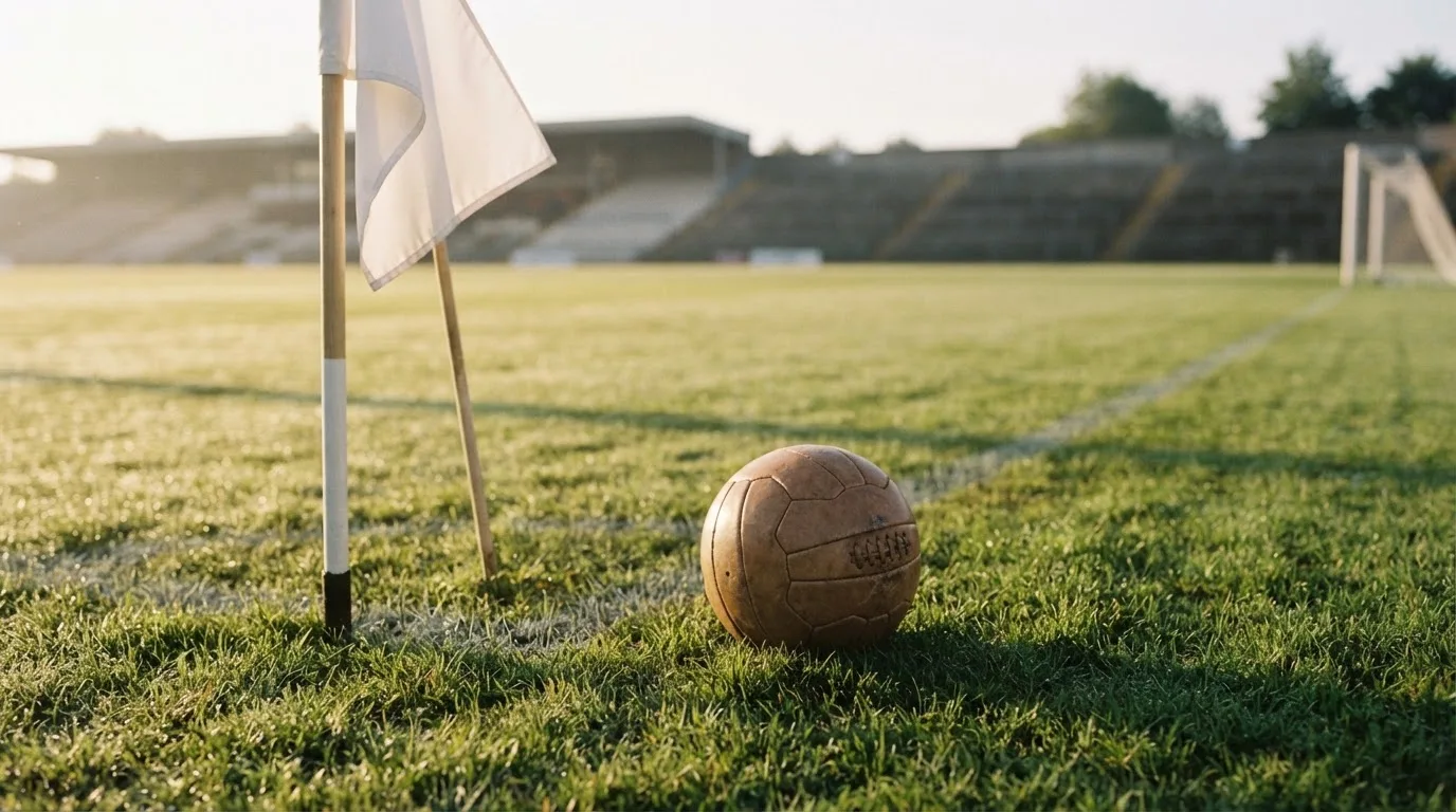 Ein Fußball liegt auf Rasen neben einer Eckfahne ohne Aufdruck auf einem leeren Platz im Morgenlicht.