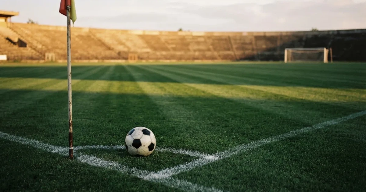 Ein Fußball liegt am Viertelkreis an der Eckfahne eines Rasenplatzes, im Hintergrund leere Tribünen in warmem Abendlicht.