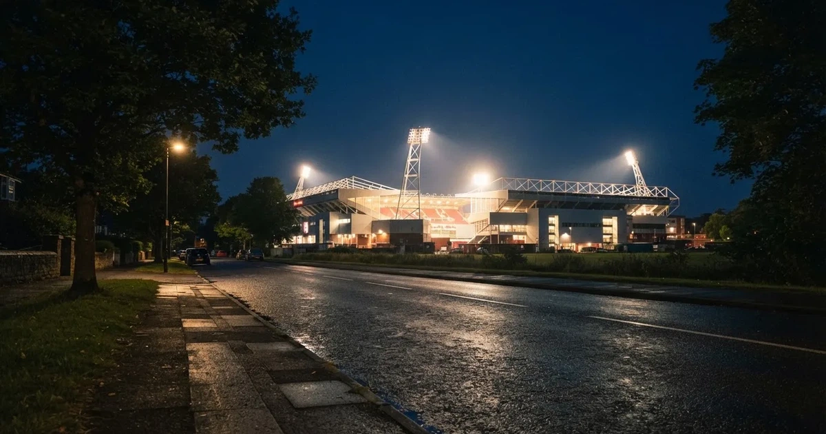 Ein hell erleuchtetes Stadion aus der Ferne bei Nacht, davor eine stille Straße und ein paar Bäume.