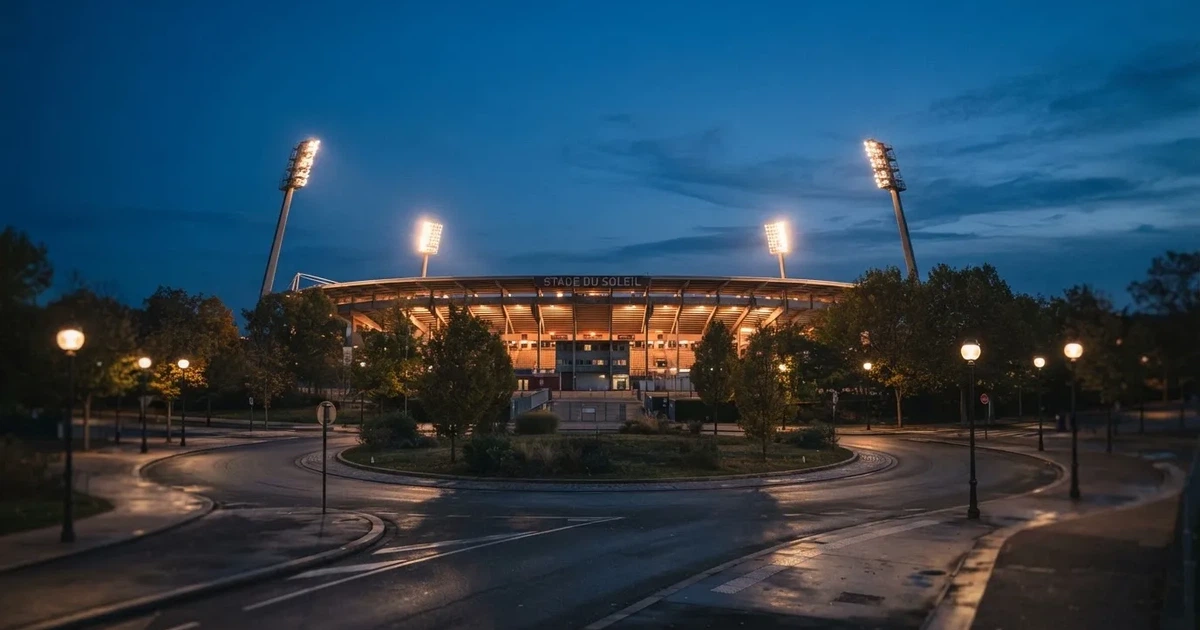Ein Fußballstadion in der Dämmerung aus der Ferne, die Flutlichter gerade eingeschaltet, davor ein Kreisverkehr mit Laternen.