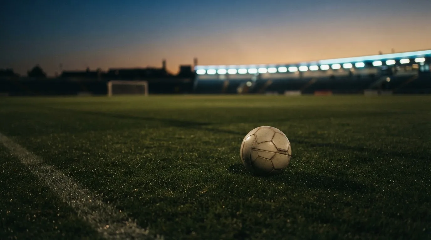 Ein Fußball liegt auf einem Rasenplatz in der Dämmerung, im Hintergrund ein ferner Stadionrand unter Flutlicht.