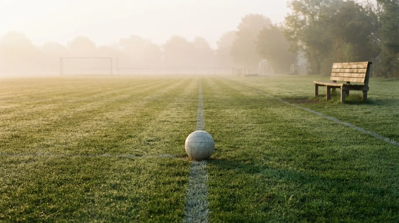 Ein Fußball liegt in der Mitte eines leeren Rasenplatzes im Morgennebel, am Rand steht eine einzelne Auswechselbank.